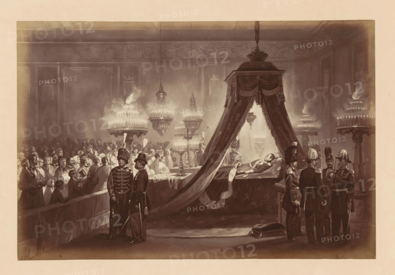 Photographic reproduction of a design of the laid-out body of King Léopold I of Belgium in the Royal Palace in Brussels, Exposition du Corps de SM Léopold 1er - au Palais de Bruxelles, photograph, reproduction, This photo is part of a cover with fourteen photos., inscription: 'Relevé de Garde.', photographer: Ghémar Frères, Brussels, after design by anonymous, c. 1861 - in or before 1866, paper, albumen print, height 168 mm x width 250 mm, Person, Leopold I (King of the Belgians), funerary ceremonial, king, Royal Palace (Brussels), Period, 1865-12, A large dimly lit interior scene shows a long room with ornate walls and high ceiling, filled with people gathered around a raised platform draped with heavy curtains. On the platform a man lies on a decorated bier beneath a canopy, dressed in formal attire and wearing medals, while attendants stand nearby in dark military uniforms with plumed helmets. Rows of seated and standing figures fill the left side of the room, many wearing head cove...