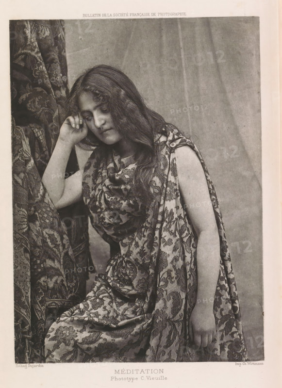 Méditation, Portrait of an unknown woman, page, photomechanical print, photographer: C. Vieuille, cliché maker: Dujardin, printer (person): Charles Wittmann, c. 1886 - in or before 1896, printer (person), paper, photoaquatint, height 170 mm x width 121 mm, adult woman, curtains, dress, gown, A black-and-white photograph of a seated young woman with long wavy hair wearing a patterned draped garment, her head tilted slightly downward and resting against one hand, the other arm hanging relaxed at her side, with patterned fabric visible behind and around her and a plain backdrop behind her.