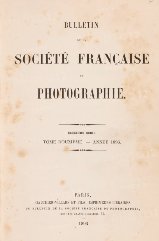 Bulletin de la Société française de photographie, book, inscription: 'Collection Jahel', publisher: Gauthier-Villars & Fils, Paris, 1896, pasteboard, biological material, paper, leather, printing, autotypie, collotype, agate marbled paper, spanish marbled paper, photoaquatint, height 251 mm x width 164 mm x thickness 38 mm, A faded, aged paper page with scattered brown foxing marks and a centered layout of large dark printed lines and decorative dividers, the top and middle areas featuring bold, wide serif word blocks and smaller lines of narrow type, with ornamental horizontal flourishes above and below a midsection, and additional smaller printed lines and blocks toward the lower area, all on a pale beige background with uneven discoloration around the edges.