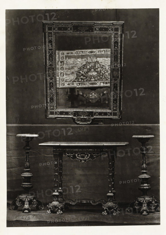 Table, two candle holders and a mirror, page, photomechanical print, name: 'De Goupil & Cie Imp. Photoglytique Paris', number: '477 478 479', photographer: Goupil & Cie., Paris, 1870, paper, Woodburytype, Woodburytypie, height 191 mm x width 134 mm, candlestick, mirror, A framed rectangular mirror with an ornate patterned border hangs on a plain dark wall, reflecting a blurred decorative textile and part of an ornamental object; below the mirror a narrow table with a carved apron and turned legs stands centered between two tall ornate candlesticks with wide circular tops and elaborately carved pedestal bases, all resting on a flat surface.