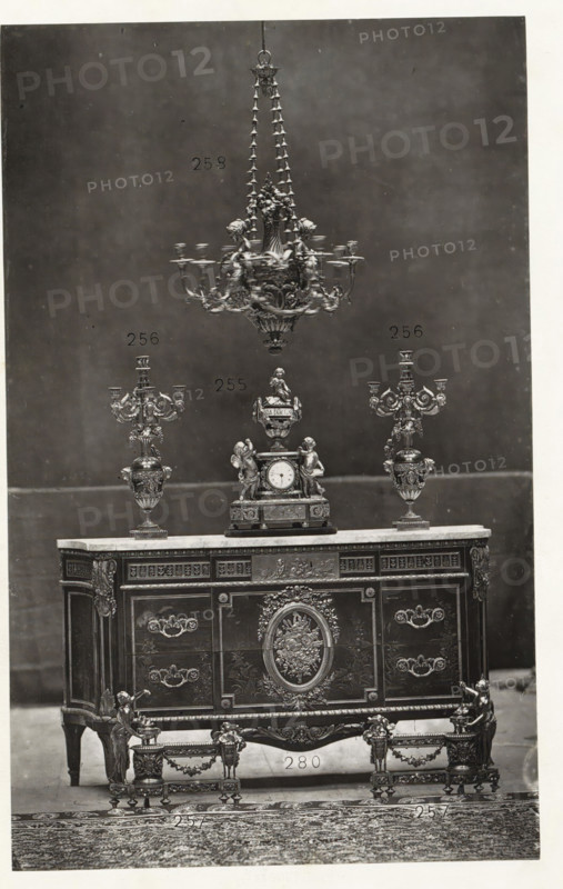 Chest of drawers with a clock and two candlesticks in Louis XVI style, page, photomechanical print, name: 'De Goupil & Cie Imp. Photoglytique Paris', number: '258 256 255 280 257', photographer: Goupil & Cie., Paris, 1870, paper, Woodburytype, Woodburytypie, height 195 mm x width 122 mm, candlestick, clock, ornamental cupboard, A tall ornate chandelier hangs from the top center, featuring multiple tiers, decorative chains, and sculpted floral and scroll details, positioned above a matching decorative clock and flanked by two elaborate candelabra with curved arms and ornamental bases. The clock sits on a carved rectangular pedestal and includes sculpted figures and ornamental vases as part of its design. All objects rest on a richly decorated wooden chest of drawers with a light-colored top, intricate paneling, carved floral medallions, decorative handles, and gilt-like embellishments on the corners and legs. In front of the chest are matching low stands with ornate legs, carved garland...