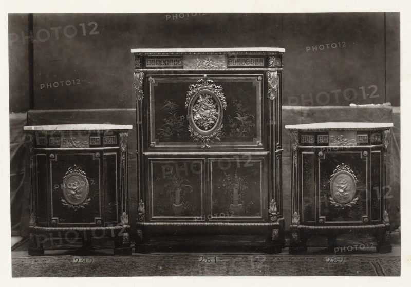 Secretary and two triangular corner pieces of furniture decorated with medallions and plant motifs, page, photomechanical print, name: 'De Goupil & Cie Imp. Photoglytique Paris', number: '281 282', photographer: Goupil & Cie., Paris, 1870, paper, Woodburytype, Woodburytypepie, height 123 mm x width 183 mm, writing-table, writing-desk, ornamental cupboard, A central ornate cabinet with a flat top sits between two smaller matching cabinets, all featuring dark wood panels with lighter tops and decorative molding; the central cabinet has an oval floral relief surrounded by carved garlands on its upper door and framed panels with painted floral motifs on its lower doors, while each side cabinet displays a similar oval relief on its front panel and decorative corner carvings, all standing on short carved legs against a plain dark backdrop with a lighter floor beneath.