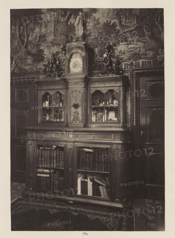 Room in the house of Antonin Terme with oak cupboard, photograph, page, Liège, c. 1880 - in or before 1885, photographer, paper, platinum process, height 223 mm x width 154 mm, art collection, art gallery, cabinet of curiosities, 'Kunst- und Wunderkammer', An ornate wooden cabinet with carved panels and glass-fronted upper compartments fills the image, the upper central section topped by a decorative clock face and a small statue, flanked by sculpted horse figures; the glass compartments display various small objects and vessels, while the lower sections reveal shelves of books and stacked papers behind glass doors; the cabinet is set against a wall covered with a detailed tapestry or mural depicting trees, foliage, and a swan in water, and parts of carved wooden doors with decorative moldings are visible at the sides, a patterned rug lies on the floor in front.