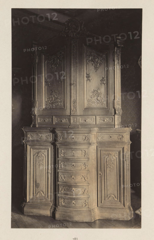 Decorated cupboard from the collection of Antonin Terme, photograph, page, Liège, c. 1880 - in or before 1885, photographer, paper, platinum process, height 224 mm x width 154 mm, art collection, art gallery, cabinet of curiosities, 'Kunst- und Wunderkammer', A tall wooden cabinet with ornate carved panels fills the frame, showing an upper section with two large doors adorned with intricate floral and scrollwork carvings and a decorative carved crest at the top, and a lower section with a central column of curved drawers flanked by paneled cupboard doors, all surfaces featuring detailed relief ornamentation and molded edges, set against a dark patterned wall and resting on a wooden floor.