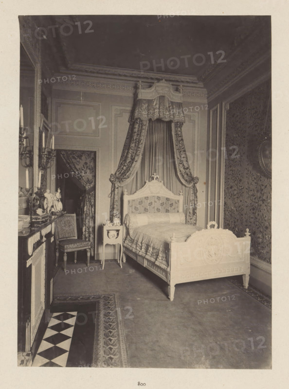 Room in the house of Antonin Terme with decorated bed, photograph, page, Liège, c. 1880 - in or before 1885, photographer, paper, platinum process, height 223 mm x width 161 mm, bed, art collection, art gallery, A photograph of an ornate bedroom showing a carved wooden bed with a decorative headboard and footboard, dressed with patterned bedding and a cylindrical bolster, positioned beneath a fabric canopy with heavy draped curtains tied back at the sides; beside the bed is a small painted bedside table holding decorative items and a chair with upholstered seat and carved legs sits near a mantelpiece topped with candlesticks and ornamental figurines; walls feature molded panels and patterned wallpaper, an ornate ceiling cornice runs along the top, and a patterned rug and a tiled border cover the floor.