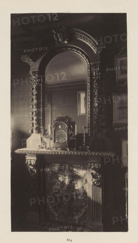 Chimney with clock in the house of Antonin Terme, photograph, page, Liège, c. 1880 - in or before 1885, photographer, paper, platinum process, height 217 mm x width 116 mm, clock, art collection, art gallery, chimney-piece, mantel piece, A tall ornate mirror with a carved arch and decorative molding sits above a mantelpiece, the mirror reflecting part of a patterned wall and a framed picture. On the mantel rests an elaborate clock with a domed top and a round face flanked by a pair of candlesticks, and the mantel edge features a carved rope-like trim. The fireplace surround displays intricate carved columns and floral or foliate designs on the panel below the mantel, and the surrounding wall shows patterned wallpaper and framed artwork. The overall tones are dark and the image appears monochrome.