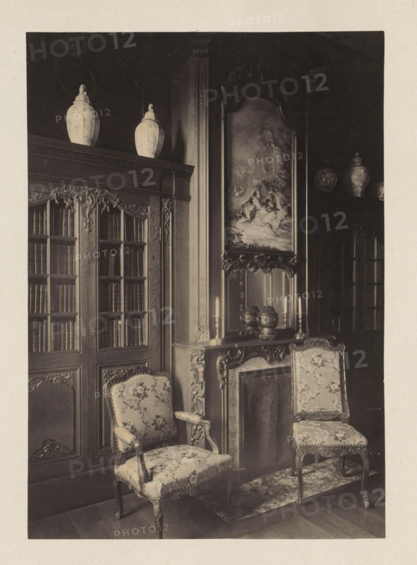 Room with chimney and two bookcases in the house of Antonin Terme, photograph, page, Liège, c. 1880 - in or before 1885, photographer, paper, platinum process, height 208 mm x width 147 mm, art collection, art gallery, chimney-piece, mantelpiece, bookcase, with doors, A sepia photograph showing an ornate interior corner with carved wooden cabinetry and a tall bookcase with glass-paneled doors filled with books, fluted decorative columns, and sculpted floral motifs; two large lidded ceramic urns sit on top of the cabinetry; a carved mantel with decorative vases and tall candlesticks stands beneath a framed painting depicting a pastoral scene; two upholstered chairs with floral fabric and carved wooden frames are positioned near the mantel on a patterned rug; additional decorative ceramic vessels are visible on a high shelf in the darker background.