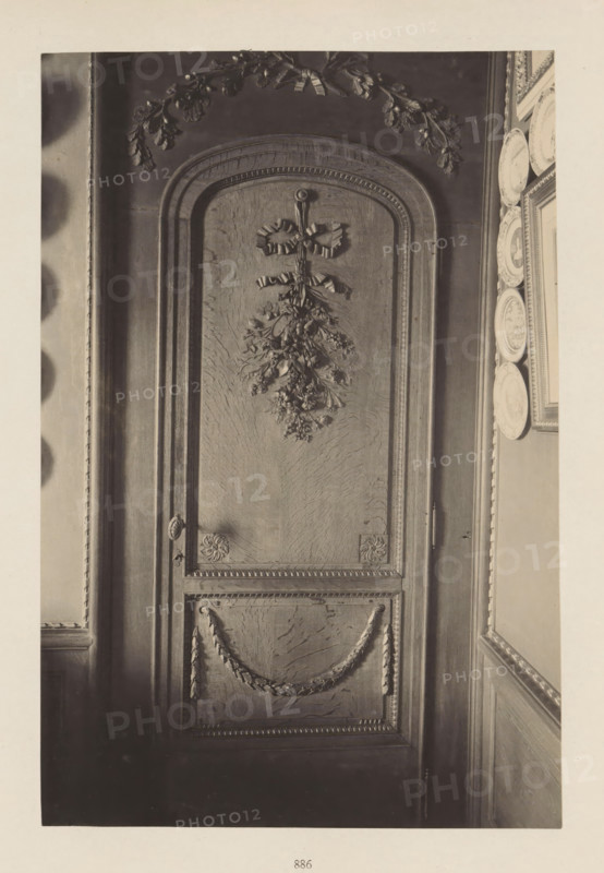 Wooden decorated door in the house of Antonin Terme, photograph, page, Liège, c. 1880 - in or before 1885, photographer, paper, platinum process, height 222 mm x width 150 mm, door, art collection, art gallery, sculpture (relief, sculpture), A tall wooden door with an arched top occupies the center of the image, featuring carved decorative elements including a ribbon-tied floral swag near the top and a carved garland draped across the lower panel, framed by beaded molding and corner rosettes; to the right of the door a narrow wall section displays a vertical row of decorative plates mounted beside a framed picture, and to the left a portion of an adjacent wall and molding is visible, all shown in a monochrome tone.