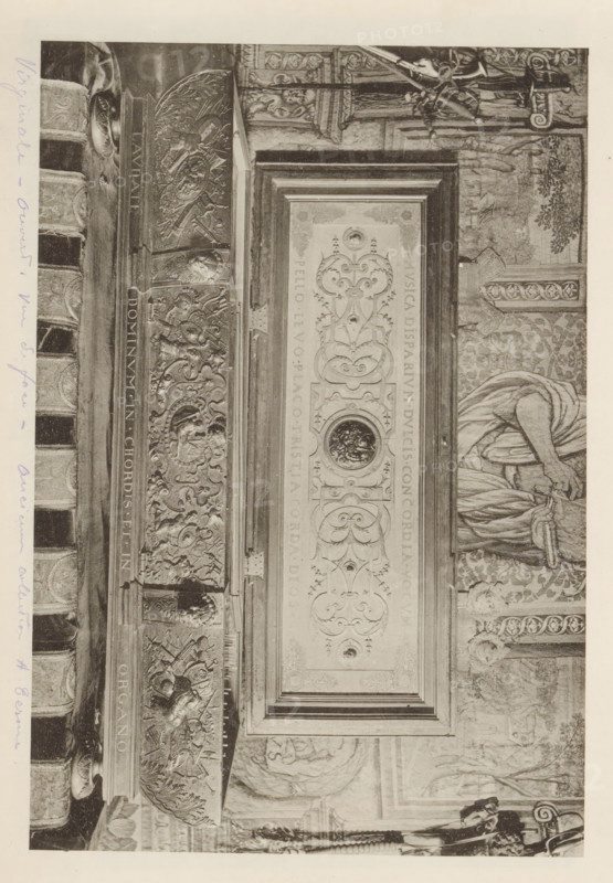 Virginal Duke of Cleves from the collection of Antonin Terme, page, photomechanical print, annotation: 'Virginale - ouvert vue ... collection A. Terme, 128 =', c. 1880 - in or before 1885, paper, collotype, photoprint, height 137 mm x width 203 mm, Print is loose in the book., spinet, virginal, art collection, art gallery, A grayscale photograph of an ornate carved doorway or panel rotated sideways, featuring an elaborate rectangular central panel with symmetrical scrollwork, circular medallions, and decorative relief elements, flanked by richly carved vertical borders with sculpted figures, faces, and heraldic motifs; to the right of the central panel part of a draped figure holding fabric is visible against a patterned wall, and above and below are architectural details including columns, sculpted friezes, and decorative moldings, with a narrow margin of blank pale paper around the image.