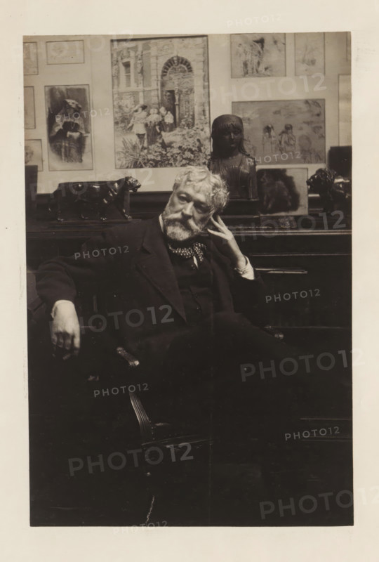 Portrait of Antonin Terme, photograph, page, c. 1880 - in or before 1885, photographer, paper, platinum process, height 186 mm x width 122 mm, Person, Antonin Terme, historical persons, An older man with a beard and mustache sits in a dark suit and patterned neckerchief, leaning his head on one hand and resting the other arm on the chair, with a contemplative expression; behind him a wooden surface holds several sculptures including a dark bust of a girl and a dark animal figure, and the wall behind displays framed artworks and illustrations in various sizes.