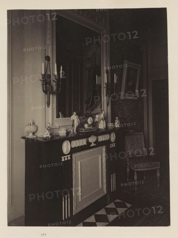 Room in the house of Antonin Terme with its collection of antiquities, statue of Hercules, clock and candlesticks, photograph, page, Liège, c. 1880 - in or before 1885, photographer, paper, platinum process, height 220 mm x width 165 mm, interior of the house, art collection, art gallery, A dark mantel with decorative panels and medallion reliefs holds various small porcelain and sculpted figurines and a central clock, flanked by two ornate wall sconces with candles; above the mantel is a large framed mirror with an embellished frame reflecting part of the room, and to the right a framed picture is hung at an angle above a patterned chair with carved legs; the floor in front of the mantel shows a checkerboard tile strip and the surrounding walls feature moulding and patterned wallpaper.