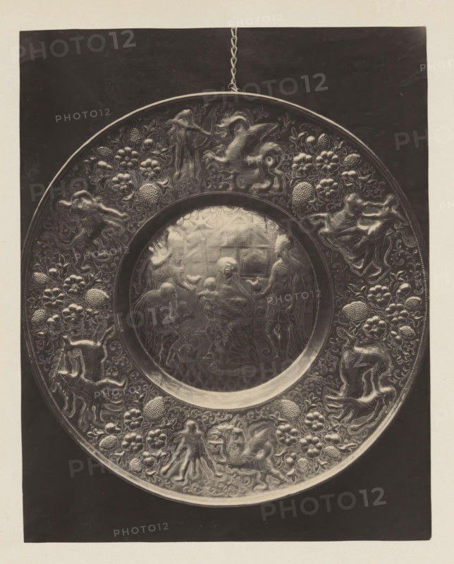 Copper bowl from the collection of Antonin Terme, photograph, page, Liège, c. 1880 - in or before 1885, photographer, paper, platinum process, height 205 mm x width 163 mm, dish, plate, saucer, art collection, art gallery, birth and youth of Christ, A round metallic plate with a raised rim hangs by a short chain against a dark rectangular background, the rim and outer band decorated with intricate reliefs of floral clusters, rounded textured fruits, and various stylized animals and humanlike figures in dynamic poses; the central circular panel contains a finely detailed scene showing several humanlike figures gathered around a seated figure holding a small child, with other standing and kneeling figures nearby and architectural and decorative motifs in the background.