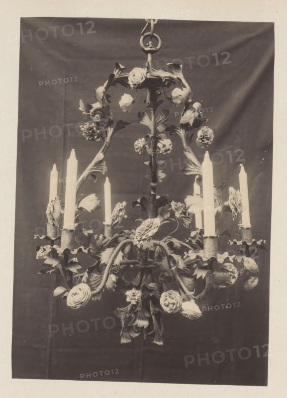 Wrought iron chandelier from the collection of Antonin Terme, photograph, page, Liège, c. 1880 - in or before 1885, photographer, paper, platinum process, height 214 mm x width 221 mm, chandelier, candelabrum, art collection, art gallery, A sepia-toned photograph of an ornate chandelier suspended from a circular hook, featuring curved metal arms decorated with sculpted leaves and numerous rose-like floral elements, with tapered candles set in scalloped candle holders distributed around the fixture; the chandelier hangs against a dark draped backdrop with visible folds and a lighter border framing the image.