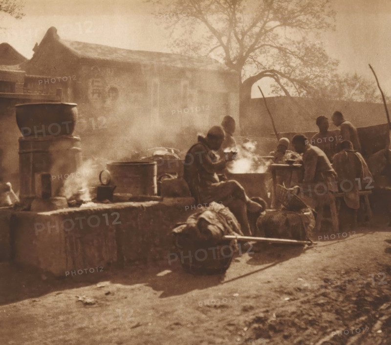 Unknown eating men, page, photomechanical print, photographer: Donald Mennie, Beijing, cliché maker: anonymous, c. 1915 - in or before 1920, paper, rotogravure, height 217 mm x width 239 mm, historical persons, eating, A sepia-toned outdoor scene showing a group of people gathered around steaming pots and cooking fires near a low stone wall, with large metal cauldrons and barrels placed on and near the wall; some figures are seated while others stand or lean, several are engaged with the steaming pots, and a person is carrying a load on a pole; in the background there is a building with a tiled roof and decorative gables, a tall tree with branches spreading overhead, and a curved archway set into a wall, all seen through hazy smoke.