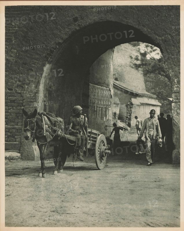 Unknown persons with horse and cart at a gate, page, photomechanical print, photographer: Donald Mennie, Beijing, cliché maker: anonymous, c. 1915 - in or before 1920, paper, rotogravure, height 252 mm x width 203 mm, historical persons, two-wheeled, animal-drawn vehicle, gate, entrance, A wide stone archway framed by textured brickwork opens onto a shadowed passage where a cart hitched to a donkey stands in front of the arch, the donkey facing forward with visible harnesses and ropes, its ears prominent; a partially shirtless figure sits on the cart holding a pole and wearing a cloth wrapped around the head; behind and within the arch a person balances a carrying pole across the shoulders with buckets hanging from its ends; another man walks forward carrying items in each hand and wearing light clothing; additional silhouetted figures stand further back in the passage; architectural details of worn walls, a wooden gate, and stone structures are visible through the arch, and a tree can...