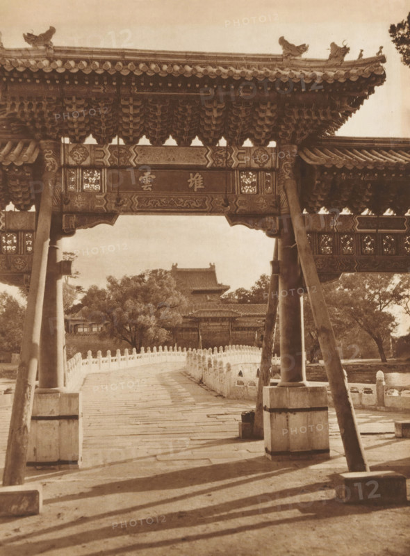 Gate and bridge at the Beihai Park in Beijing, page, photomechanical print, photographer: Donald Mennie, Beijing, cliché maker: anonymous, c. 1915 - in or before 1920, paper, rotogravure, height 274 mm x width 204 mm, bridge, gate, entrance, Beihai Park, A sepia-toned photograph shows a large traditional wooden gate with ornate carved beams and tiled eaves supported by thick pillars and angled wooden braces, framing a stone bridge with a balustrade of repeating short posts that leads toward a distant building with layered tiled roofs partially obscured by trees; decorative patterns appear on the gate beams and the roof edges, and shadows from the structure and posts stretch across the stone pavement in the foreground.
