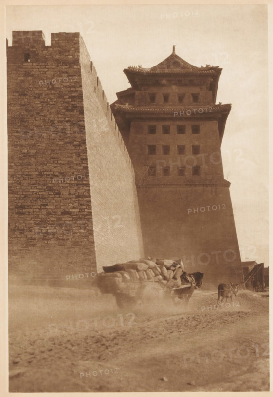 Cart pulled by a horse near a building, page, photomechanical print, photographer: Donald Mennie, Beijing, cliché maker: anonymous, c. 1915 - in or before 1920, paper, rotogravure, height 276 mm x width 191 mm, two-wheeled, animal-drawn vehicle, civic architecture, edifices, dwellings, A tall brick wall rises along the left side of the image next to a multi-tiered tower with eaves and small square openings, both rendered in a monochrome sepia tone; at the base of the structures a cart loaded with rounded sacks is being pulled by an animal, and another animal stands nearby, while dust or haze swirls around their legs and the foreground shows a textured, dusty ground.