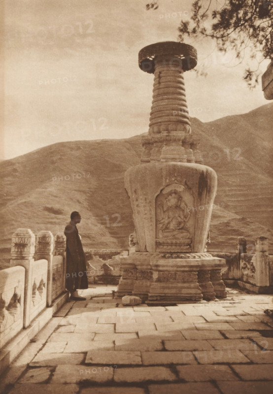 Unknown man at a stupa, page, photomechanical print, photographer: Donald Mennie, Beijing, cliché maker: anonymous, c. 1915 - in or before 1920, paper, rotogravure, height 279 mm x width 201 mm, adult man, statues, paintings, etc., objects of worship (non-Christian religions), Temple of Azure Clouds, A sepia-toned photograph shows a stone terrace with a tiled floor and carved balustrade, a large carved stupa-like monument occupying the right foreground with ornate relief sculpture on its rounded base and a fluted column rising to a wide circular capital, a solitary robed figure standing near the balustrade facing the monument, distant terraced hills forming the background, and tree branches extending into the upper right corner.