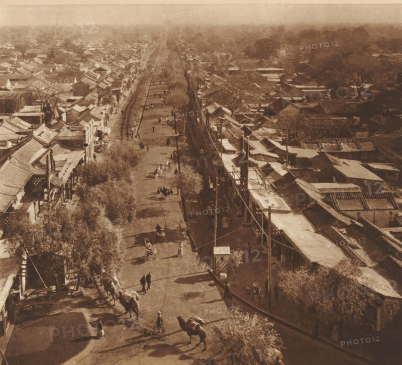 Street view in Beijing, page, photomechanical print, photographer: Donald Mennie, Beijing, cliché maker: anonymous, c. 1915 - in or before 1920, paper, rotogravure, height 218 mm x width 237 mm, street, A high vantage street view shows a long straight road flanked by rows of low buildings with sloping tiled roofs and awnings, trees lining parts of the street, overhead utility poles and wires along one side, and groups of people and animal-drawn carts moving along the roadway; the scene is captured in a sepia tone with distant rooftops and treetops fading into a hazy background.