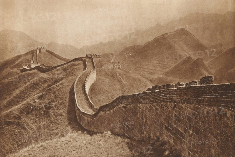 Great Wall of China, page, photomechanical print, photographer: Donald Mennie, Beijing, cliché maker: anonymous, c. 1915 - in or before 1920, paper, rotogravure, height 194 mm x width 280 mm, walls, fortified city, A long stone wall with battlements winds along a series of rolling, grassy hills receding into misty mountains, with several watchtowers and fortified structures punctuating the wall; the scene is rendered in a sepia tone, showing textured stonework on the wall faces, paths along the wall, undulating slopes, and distant layered ridgelines fading into the background.
