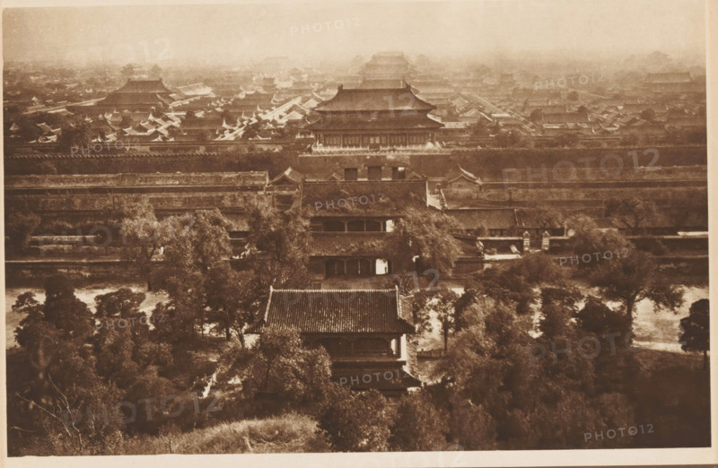 View of the Forbidden City in Beijing, page, photomechanical print, photographer: Donald Mennie, Beijing, cliché maker: anonymous, c. 1915 - in or before 1920, paper, rotogravure, height 182 mm x width 276 mm, city-view in general, 'veduta', Forbidden City, A sepia-toned aerial view shows a dense cluster of traditional tiled-roof buildings and pavilions arranged behind multiple long horizontal walls, with large roofed halls rising toward the background and rows of smaller peaked roofs filling the midground; trees and leafy vegetation appear in the foreground interspersed with pathways and courtyard areas, while the distant upper portion of the scene is hazy and shows further layered rooftops fading into the background.