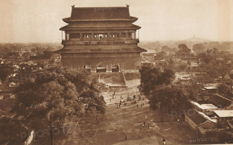 Gu Lou in Beijing, page, photomechanical print, photographer: Donald Mennie, Beijing, cliché maker: anonymous, c. 1915 - in or before 1920, paper, rotogravure, height 183 mm x width 279 mm, landscape with tower or castle, Gu Lou, A large multi-tiered traditional building with sweeping tiled roofs stands centrally, elevated on a wide base with arched openings at its ground level and an open gallery on the upper story. In front of the building a broad dirt pathway slopes downward through an open area where small groups of people are scattered, some standing and some walking. Trees with dense foliage frame the foreground and sides, and low rooftops and clustered buildings extend outward behind and beside the central structure. The image has a sepia tone and a hazy distant horizon with indistinct shapes of more trees and structures.