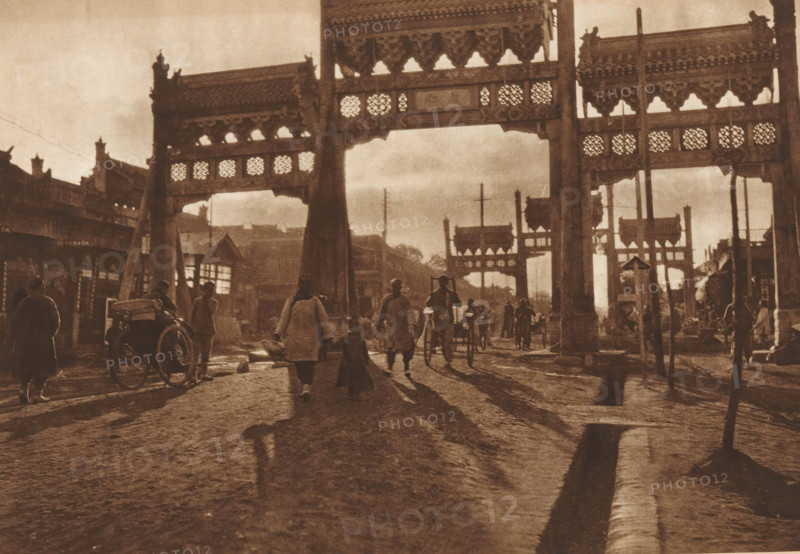 Paifang in Beijing, page, photomechanical print, photographer: Donald Mennie, Beijing, cliché maker: anonymous, c. 1915 - in or before 1920, paper, rotogravure, height 198 mm x width 278 mm, gate, entrance, A wide street scene in a sepia tone shows large ornate gate structures with carved lattice panels spanning the roadway, casting shadows on the uneven ground. People walk and stand beneath and around the gates, some wearing long coats and carrying items, others pulling or riding simple rickshaws. Shadows from the figures stretch along the cobblestone surface toward the foreground. Flanking buildings with pitched roofs and decorative eaves line the sides of the street, and utility poles rise between the gate openings. The overall lighting is low and diffuse, creating a muted, aged appearance.