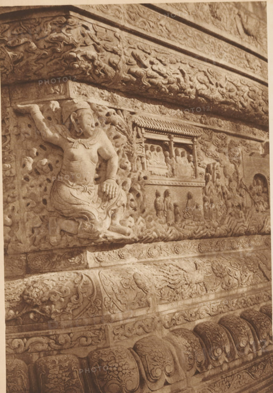 Detail of a stupa, page, photomechanical print, photographer: Donald Mennie, Beijing, cliché maker: anonymous, c. 1915 - in or before 1920, paper, rotogravure, height 279 mm x width 196 mm, statues, paintings, etc., objects of worship (non-Christian religions), A close view of an ornate carved stone relief showing a seated figure with one arm raised and the other resting on a knee, surrounded by swirling cloud-like and floral patterns, with a rectangular niche containing smaller seated figures and standing attendants, bordered by decorative bands and layered carved moldings, all rendered in a monochrome sepia tone.