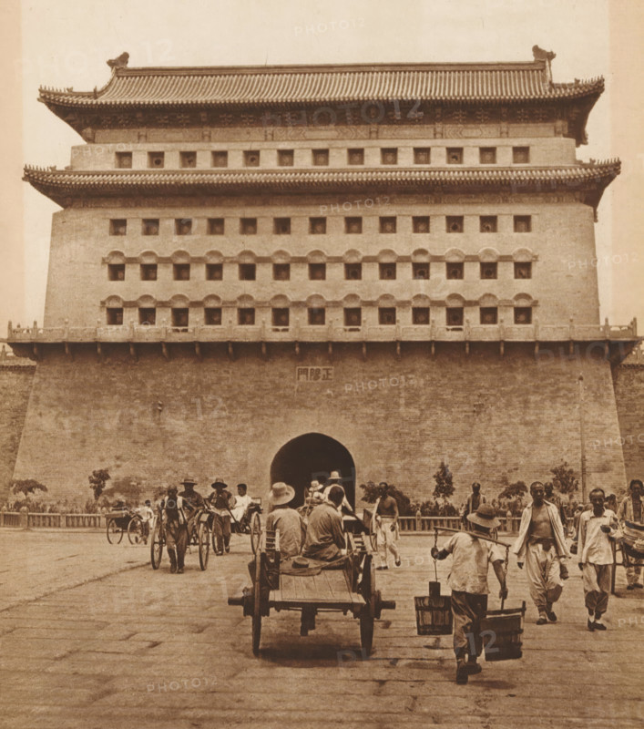 View of Zhengyang Men, page, photomechanical print, photographer: Donald Mennie, Beijing, cliché maker: anonymous, c. 1915 - in or before 1920, paper, rotogravure, height 245 mm x width 216 mm, gate, entrance, Zhengyang Men, A large multi-tiered stone and brick gatehouse with tiled overhanging roofs and rows of small square openings dominates the upper part of the image, while a wide arched passage sits at its center; in front of the gatehouse a paved plaza shows numerous people walking and working, some pulling or riding in two-wheeled rickshaws and carts, one cart with two seated figures viewed from behind, others carrying buckets suspended from a shoulder pole, and a few standing or walking individuals wearing hats and loose clothing.