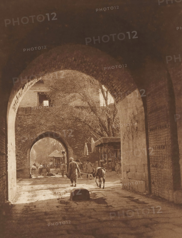 View of two gates in Beijing, page, photomechanical print, photographer: Donald Mennie, Beijing, cliché maker: anonymous, c. 1915 - in or before 1920, paper, rotogravure, height 257 mm x width 203 mm, gate, entrance, A sepia-toned view through a series of brick archways opening onto a street scene, with the nearest archway framing a passageway floor showing wet patches and a stone in the foreground. Beyond the arch, a person walks away leading a donkey on a rope, other figures and animals are visible further down the street, and a small wooden shelter or stall stands to one side. Trees with bare branches rise above the far wall, and the brickwork of the arches and adjoining walls shows texture and wear. Light filters through the arches creating areas of shadow and brightness along the ground and walls.