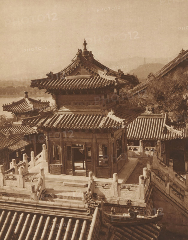 Summer Palace in Beijing, page, photomechanical print, photographer: Donald Mennie, Beijing, cliché maker: anonymous, c. 1915 - in or before 1920, paper, rotogravure, height 258 mm x width 202 mm, palace, Summer Palace, A sepia-toned photograph showing traditional East Asian architecture with layered tiled roofs, ornate ridge decorations, and carved eaves; a central wooden pavilion with open doorways and lattice panels sits on a raised stone terrace surrounded by balustrades and stone stairways; surrounding buildings with similar tiled roofs and decorative roof figures are visible, along with a hillside and distant landscape in the background.