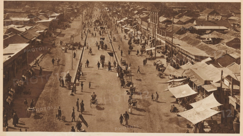 Street view in Beijing, page, photomechanical print, photographer: Donald Mennie, Beijing, cliché maker: anonymous, c. 1915 - in or before 1920, paper, rotogravure, height 164 mm x width 290 mm, street, A sepia-toned aerial view of a wide street lined with low buildings and awnings on both sides, with a raised central divider and lamp posts running down its length, showing many people walking or standing, several handcarts and rickshaws moving along the roadway, and groups of figures gathered near the storefronts and under fabric canopies, with tiled roofs extending into the distance and long shadows cast across the ground.