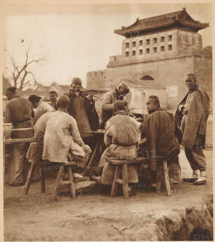 Group of unknown men, some of whom smoke pipes, page, photomechanical print, photographer: Donald Mennie, Beijing, cliché maker: anonymous, c. 1915 - in or before 1920, paper, rotogravure, height 242 mm x width 217 mm, historical persons, adult man, pipe, tobacco, A sepia-toned scene shows a group of people gathered around a low wooden table with simple stools, most seated with their backs or sides to the camera and wearing layered, textured clothing and wrapped garments. Two older men stand near the table, one playing a long pipe and another leaning over a large pot or basin while holding a utensil. A tall man stands to the right wearing a long quilted coat and looking toward the camera. In the background a multi-tiered brick building with a tiled roof and rows of small square openings rises above the group, and bare tree branches frame the left side of the image. The ground in the foreground is bare earth with scattered small debris and rough stone edging visible at the bottom right.