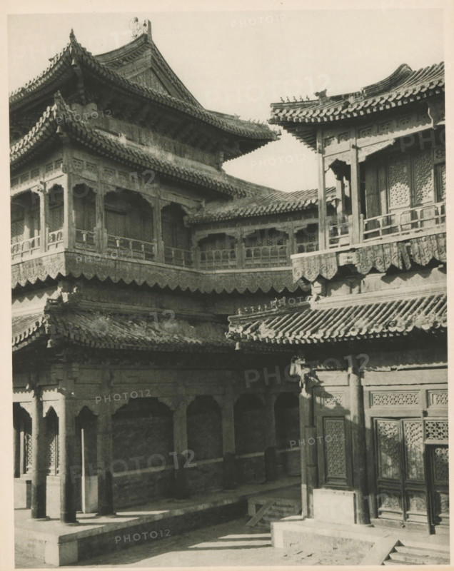 Yonghe Temple in Beijing, page, photomechanical print, photographer: Donald Mennie, Beijing, cliché maker: anonymous, c. 1915 - in or before 1920, paper, rotogravure, height 205 mm x width 258 mm, temple, shrine, non-Christian religions, A sepia-toned photograph of a traditional multi-tiered building with ornate upturned eaves and tiled roofs, showing carved wooden brackets and latticework panels. Balconies with carved railings run along the upper levels, supported by columns with decorative capitals. Scalloped wooden friezes hang beneath the rooflines, and patterned window or door panels are visible on the right. Stone steps and a raised platform lead into a shaded colonnaded area with arched openings, and sunlight casts shadows across the courtyard and lower walls.