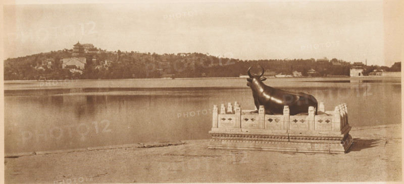 View of the Winter Palace in Beijing, page, photomechanical print, photographer: Donald Mennie, Beijing, cliché maker: anonymous, c. 1915 - in or before 1920, paper, rotogravure, height 178 mm x width 278 mm, trees, palace, A sepia-toned scene of a large body of water with a shoreline in the foreground and a hillside with buildings and trees across the water, with a dark, polished statue of a reclining bull placed on an ornate rectangular stone platform near the water’s edge; the platform has carved railings and decorative details, and the bull faces toward the water with curved horns and a smooth surface, while the distant shore shows pagoda-like structures and clustered foliage under a pale sky.