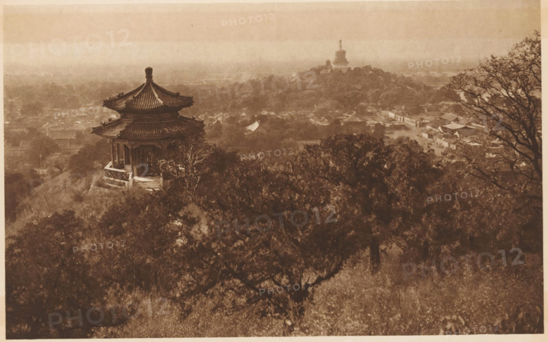 Bronze sculpture of an ox on a river near the Summer Palace in Beijing, page, photomechanical print, photographer: Donald Mennie, Beijing, cliché maker: anonymous, c. 1915 - in or before 1920, paper, rotogravure, height 138 mm x width 289 mm, sculpture, river, Summer Palace, A sepia-toned elevated view showing a traditional multi-tiered pavilion with a curved tiled roof perched on a hillside, surrounded by dense trees and foliage in the foreground, with a distant landscape of low buildings and another large stupa-like structure on a hill shrouded in haze, and a bare-branched tree framing the right edge of the scene.
