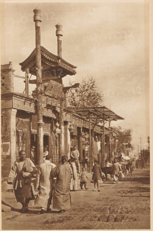 Street view in Beijing, page, photomechanical print, photographer: Donald Mennie, Beijing, cliché maker: anonymous, c. 1915 - in or before 1920, paper, rotogravure, height 279 mm x width 188 mm, street, A sepia-toned street scene shows a row of low buildings with ornate eaves and wooden columns supporting a projecting roof structure; tall carved posts rise above the roofline topped with capped finials. People in traditional loose garments walk and stand along the unpaved road, including adults and children, some carrying bundles or holding small objects. Market-style awnings and simple wooden frameworks extend from the building fronts, and hanging items are visible near doorways. A few animals are present among the people toward the middle distance. Trees rise behind the buildings and the sky appears open and mostly clear above the scene.