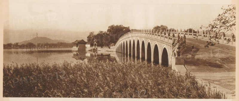 Bridge at the Summer Palace in Beijing, page, photomechanical print, photographer: Donald Mennie, Beijing, cliché maker: anonymous, c. 1915 - in or before 1920, paper, rotogravure, height 124 mm x width 289 mm, river, bridge, A sepia-toned scene shows a curved stone bridge with multiple archways reflected in calm water, ornamental balustrades lining its sides, and sculpted figures at the near end of the bridge. Tall grasses fill the foreground along the water’s edge. On the far side of the bridge a pavilion with a pitched roof and adjacent small structures sit beside trees that extend along the shoreline. Hills rise in the distant background beneath a pale sky.