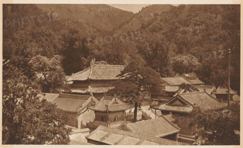 View of a landscape with mountains and roofs, page, photomechanical print, photographer: Donald Mennie, Beijing, cliché maker: anonymous, c. 1915 - in or before 1920, paper, rotogravure, height 172 mm x width 277 mm, roof (of house or building), mountains, A sepia-toned aerial view showing traditional tiled roofs and ornate eaves of a clustered complex of buildings surrounded by dense trees and hills, with layered rooftops, decorative ridgelines, a few tall trees rising among the structures, shadowed courtyards and pathways between the buildings, and a forested hillside forming the background.