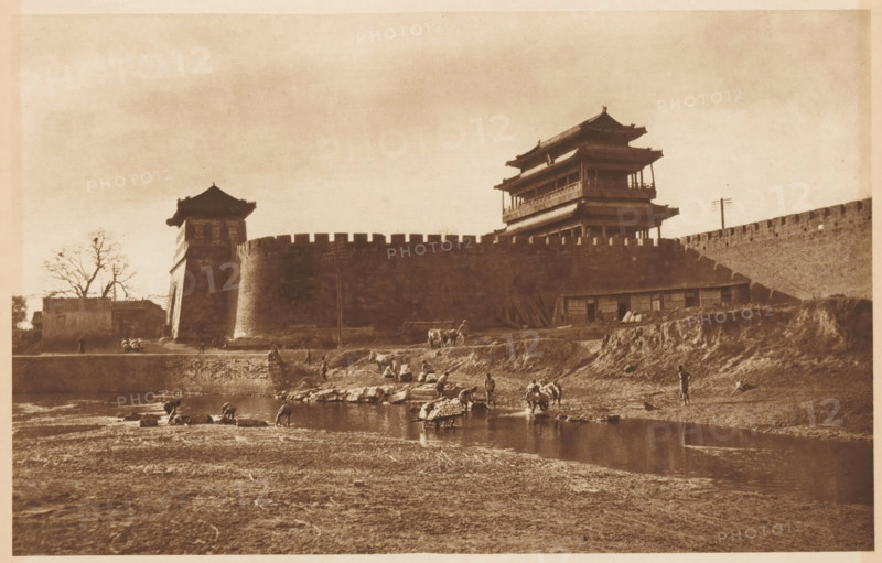 Unknown persons near a body of water along a city wall, page, photomechanical print, photographer: Donald Mennie, Beijing, cliché maker: anonymous, c. 1915 - in or before 1920, paper, rotogravure, height 179 mm x width 278 mm, historical persons, landscapes with waters, waterscapes, seascapes (in the temperate zone), city-walls, A sepia-toned scene shows a large stone wall with battlements and two multi-tiered pavilion structures rising above it, each with curved eaves. In front of the wall a sloped embankment leads down to a shallow river where several people are standing and crouching at the water's edge, some with bundles and some near small boats or rafts. Livestock and pack animals are present on the bank and near the water. On the left side a tree stands beside a smaller building adjacent to a tower incorporated into the wall. The foreground is an expanse of textured, bare ground stretching toward the river.