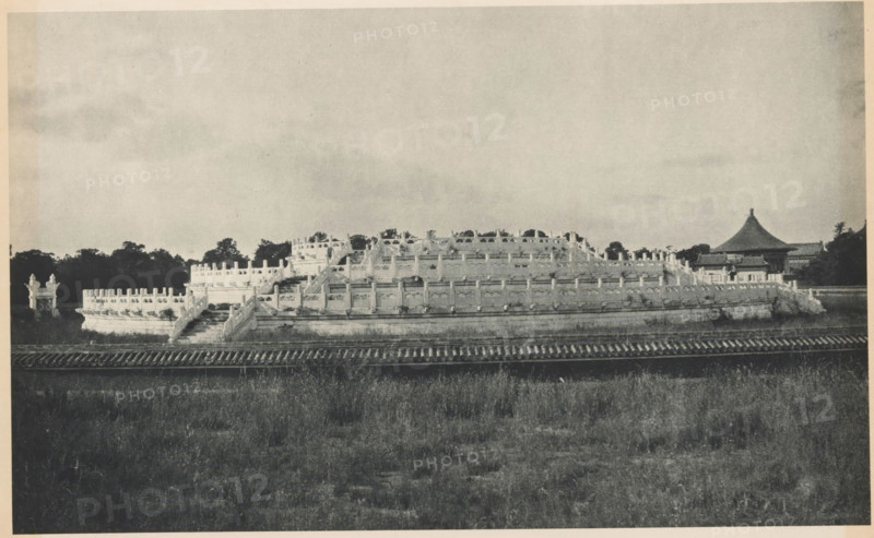 Altar of Heaven in Beijing, page, photomechanical print, photographer: Donald Mennie, Beijing, cliché maker: anonymous, c. 1915 - in or before 1920, paper, rotogravure, height 175 mm x width 280 mm, temple, shrine, non-Christian religions, Temple of Heaven, A wide black-and-white photograph of an ornate multi-tiered circular stone terrace with carved balustrades and staircases, set behind a low tiled wall and in front of a grassy foreground. The terrace features repeating decorative posts and relief panels along its tiers, with a wide central stairway leading upward. In the background there are dense dark-tree silhouettes and a pavilion-like building with a steep conical roof visible to one side. The sky above is pale and mostly empty.