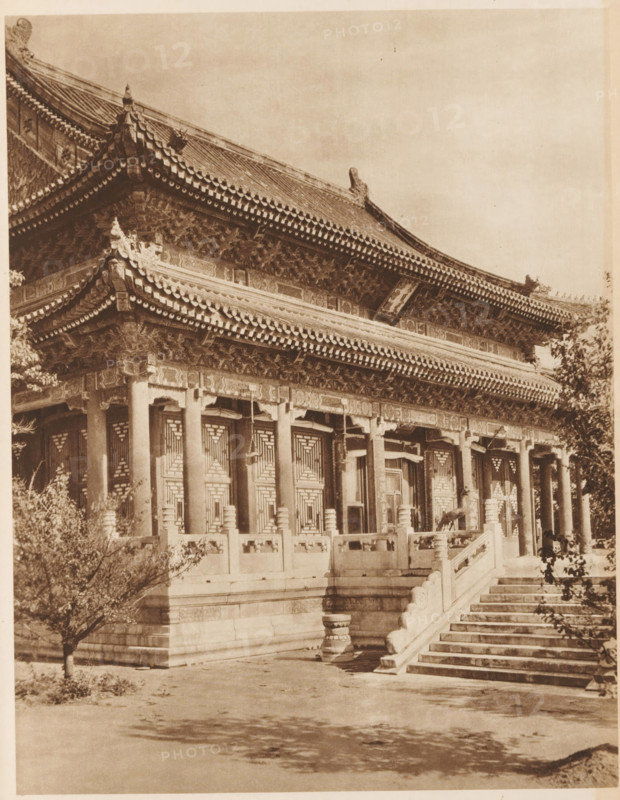 Summer Palace in Beijing, page, photomechanical print, photographer: Donald Mennie, Beijing, cliché maker: anonymous, c. 1915 - in or before 1920, paper, rotogravure, height 260 mm x width 204 mm, palace, Summer Palace, A sepia-toned photograph of a traditional multi-tiered building with ornate upturned eaves and tiled roofs, showing detailed carved brackets and decorative roof ridges. The facade features a row of tall columns supporting the overhanging eaves and intricately patterned window or door panels set between the columns. A stone balustrade runs along a raised platform in front of the building, interrupted by a wide flight of stone steps leading up to the entrance. Trees with sparse foliage frame the scene at the left and right edges, and shadowed ground stretches in the foreground.