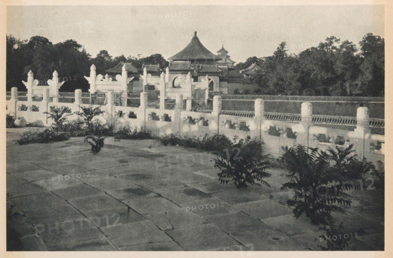 Altar of Heaven in Beijing, page, photomechanical print, photographer: Donald Mennie, Beijing, cliché maker: anonymous, c. 1915 - in or before 1920, paper, rotogravure, height 186 mm x width 278 mm, temple, shrine, traditional Chinese religions, Temple of Heaven, A wide stone courtyard with large rectangular paving stones fills the foreground, dotted with clusters of leafy plants. A low decorative balustrade with carved posts and openings runs across the middle ground, showing repeated ornamental pillars and rail sections. Behind the balustrade a series of traditional tiled-roof buildings rise, featuring curved eaves and a prominent multi-tiered roof with a pointed top. Dense trees and foliage form a dark backdrop behind the buildings under a pale sky. The image is monochrome with varied tones from light stone to dark vegetation.