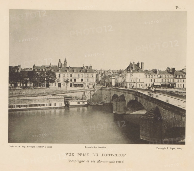 View of the Pont Neuf in Compiègne, France, Vue Prize du Pont-Neuf, Compiègne et ses Monuments (oise), page, photomechanical print, number: 'Pl. I.', inscription: 'Reproduction interdite.', maker: Jules Royer, Compiègne, maker: Augustin Boutique, Douai, c. 1890 - in or before 1895, paper, collotype, photoprint, height 121 mm x width 160 mm, Print opposite title page., city-view in general, 'veduta', bridge, A sepia-toned photograph showing a river with a stone arched bridge crossing it, a riverside building with windows and a long covered structure floating at the waterline, and a row of multi-story buildings with pitched roofs, chimneys, and storefronts lining the far bank; there are small groups of people on the bridge and along the street, trees in front of some buildings, and architectural details like a tower and spires visible above the rooftops.
