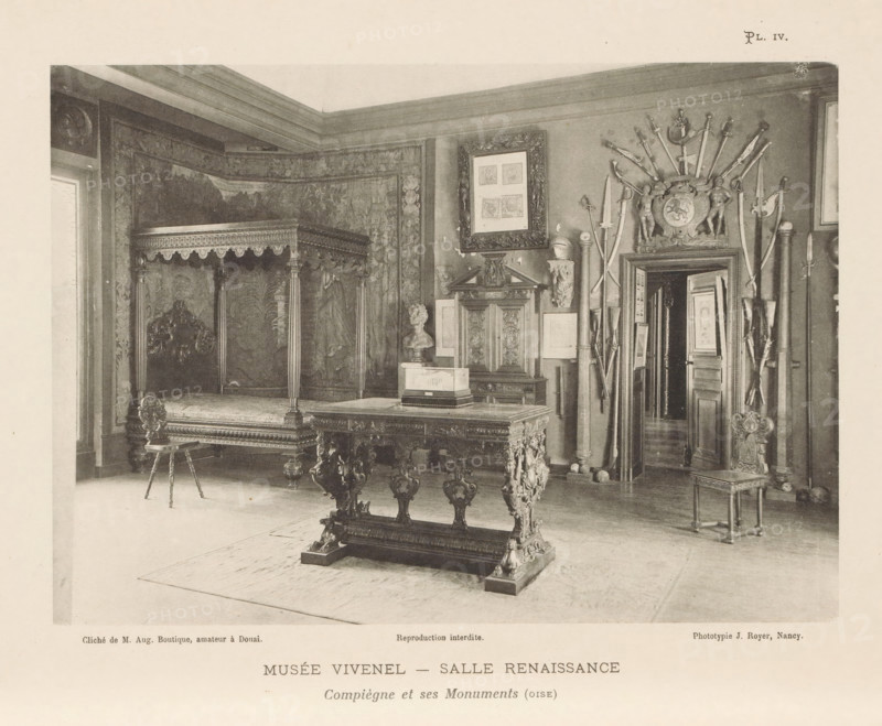 View of a room with Renaissance art in the Musée Antoine Vivenel in Compiègne, France, Musée Vivenel - Salle Renaissance, Compiègne et ses Monuments (oise), page, photomechanical print, inscription: 'Reproduction interdite.', number: 'Pl. IV.', maker: Jules Royer, Compiègne, maker: Augustin Boutique, Douai, c. 1890 - in or before 1895, paper, collotype, photoprint, height 123 mm x width 172 mm, Print on second unnumbered page after page 4., permanent exhibition, museum, furniture and household effects, A photograph of an interior room showing ornate antique furniture and wall decorations, including a large canopy bed with carved posts and a detailed headboard partially covered by patterned tapestries, a heavy carved table with ornate legs in the foreground, a small decorative stool with turned legs, and a chair with a carved back at the right; on the right wall there is an arrangement of swords and polearms fanning out above a decorative crest and a doorway that reveals another room wi...