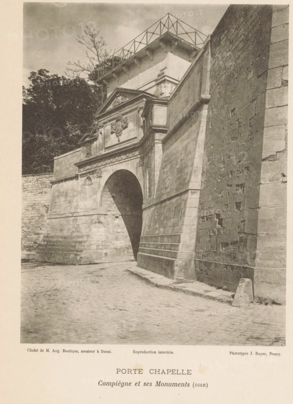 View of a chapel door in Compiègne, France, Porte Chapelle, Compiègne et ses Monuments (oise), page, photomechanical print, number: 'Pl. VIII.', inscription: 'Reproduction interdite.', maker: Jules Royer, Compiègne, maker: Augustin Boutique, Douai, c. 1890 - in or before 1895, paper, collotype, photoprint, height 160 mm x width 124 mm, church (exterior), by, A sepia-toned photograph shows a stone archway set into a large masonry wall with a sloping buttress beside it; above the arch is an ornate pediment with sculpted decoration and moulded cornices, and a heavy stone façade with architectural details. The cobbled ground in front of the arch slopes slightly and meets the base of the wall. To the left of the arch a lower stone wall and dense foliage with tall trees are visible, and a bare-branched tree rises behind the structure. The right side of the image is dominated by a tall, textured stone wall with visible joints and weathering, and the upper portion of the scene shows a flat-roo...
