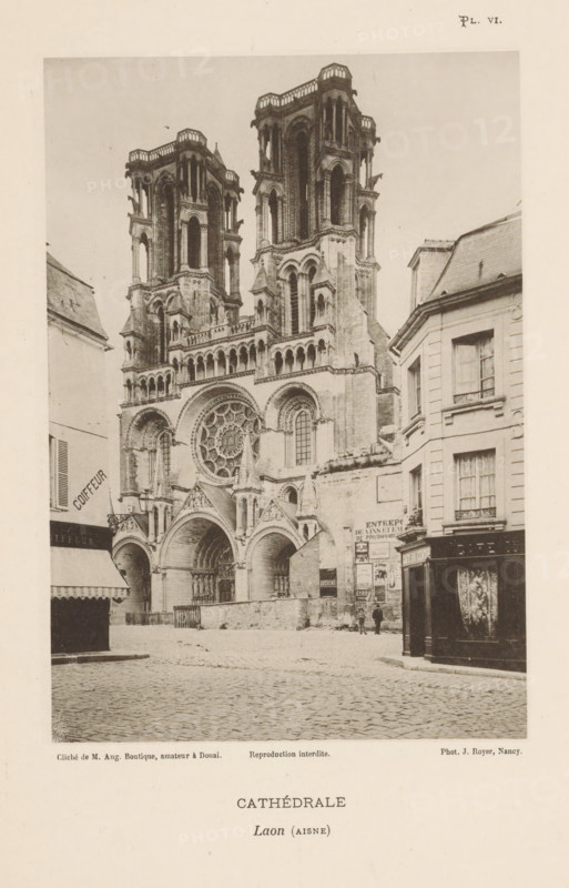 View of the Cathedral of Laon, France, Cathédrale, Laon (Aisne), page, photomechanical print, inscription: 'Reproduction interdite.', number: 'Pl. VI.', maker: Jules Royer, maker: Augustin Boutique, Douai, c. 1891 - in or before 1896, paper, collotype, photoprint, height 161 mm x width 112 mm, Print on fifth unnumbered page after page 4., church (exterior), Laon Cathedral, A sepia-toned photograph showing a large Gothic cathedral façade with two tall, ornate towers rising above a detailed front including arched doorways, a large circular rose window, decorative columns, and sculpted elements; cobblestone street in front of the cathedral leads past adjacent multi-story buildings with shuttered windows and shopfronts, one with an awning and another with a dark wooden paneled exterior; a few small figures stand near the base of the cathedral by a wall plastered with posters; the sky appears plain and light in tone above the scene.