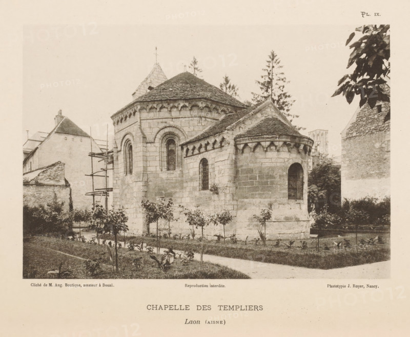 View of the Templar Chapel in Laon, France, Chapelle des Templiers, Laon (Aisne), page, photomechanical print, number: 'Pl. IX.', inscription: 'Reproduction interdite.', maker: Jules Royer, maker: Augustin Boutique, Douai, c. 1891 - in or before 1896, paper, collotype, photoprint, height 118 mm x width 171 mm, Print on third unnumbered page after page 6., parts of church exterior and annexes: chapel - QQ - small church, chapel, A sepia-toned photograph of a stone chapel with rounded apses and steep tiled roofs, showing carved corbels under the eaves and narrow arched windows with dark panes, surrounded by a grassy foreground with small plants and a narrow path, adjacent stone and plaster buildings with scaffolding visible on one side, tall trees rising behind the chapel, and leafy branches intruding from the top right edge of the image.