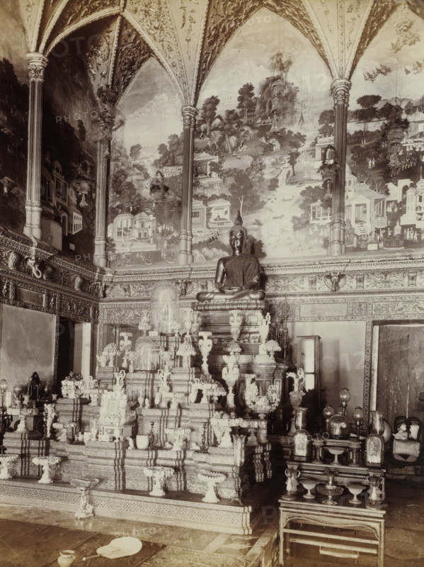 Interior of the Wat Pra Keo Temple (Temple of the Emerald Buddha) in Bangkok, album page, photograph, Part of Album with 50 photos of a trip through Southeast Asia., check stamp: 'GR Lambert Singapore', photographer: GR Lambert & Co., Bangkok, c. 1870 - c. 1900, paper, albumen print, height 273 mm x width 210 mm, temple, shrine, Hinduism, Buddhism, Jainism, A large seated statue on a raised ornate platform sits centrally beneath a vaulted ceiling with decorative arches and painted mural panels depicting trees, buildings, and landscape scenes, flanked by tall carved columns; the platform and surrounding area are filled with multiple intricately carved pedestals, vases, urns, and decorative objects arranged in tiers, and smaller altarpieces and covered displays occupy the foreground and sides, with patterned wall friezes and ornate molding running along the room, and a low mat and small vessels visible on the floor in front.