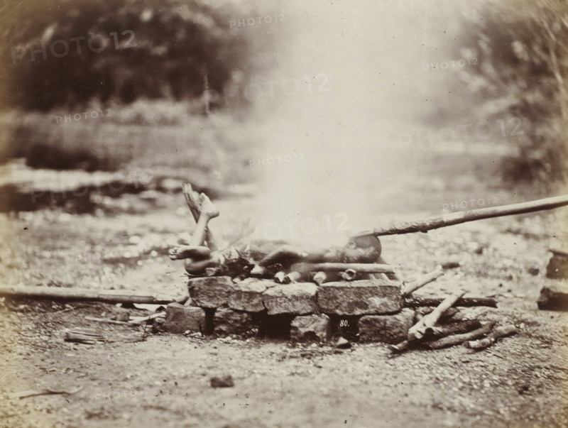 Cremation of the body of a boy in Siam, album page, photograph, Part of Album with 50 photos of a trip through Southeast Asia., negative number: '80', check stamp: 'GR Lambert & Co Singapore', photographer: GR Lambert & Co., Thailand, c. 1870 - c. 1900, paper, albumen print, height 209 mm x width 275 mm, body-burning, cremation, A sepia-toned photograph showing a rocky ground with a low stone platform in the center foreground, on top of which a human figure lies stretched out with legs raised and crossed at the ankles and arms extended alongside the body, a plume of pale smoke or steam rising vertically from the middle of the platform and obscuring the torso, a long wooden pole resting near the head and extending toward the right edge of the image, scattered logs and sticks around the platform, and a blurred natural background of foliage and uneven terrain.